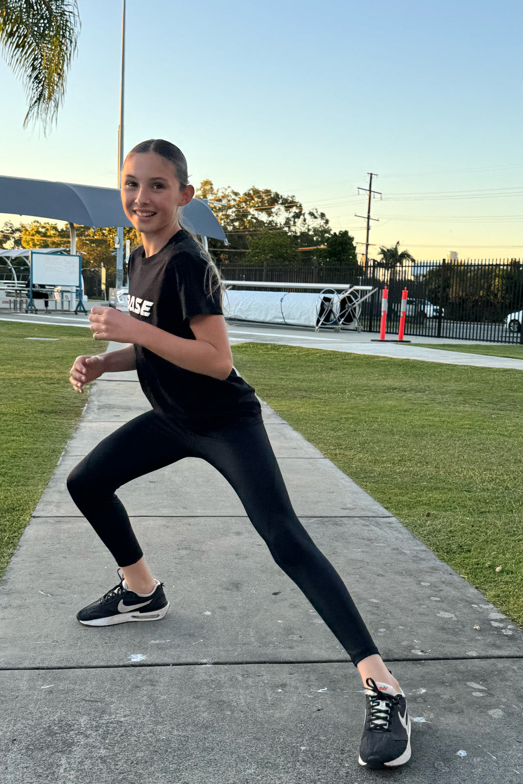 Person in black athletic wear standing on a sidewalk with grass and trees in the background