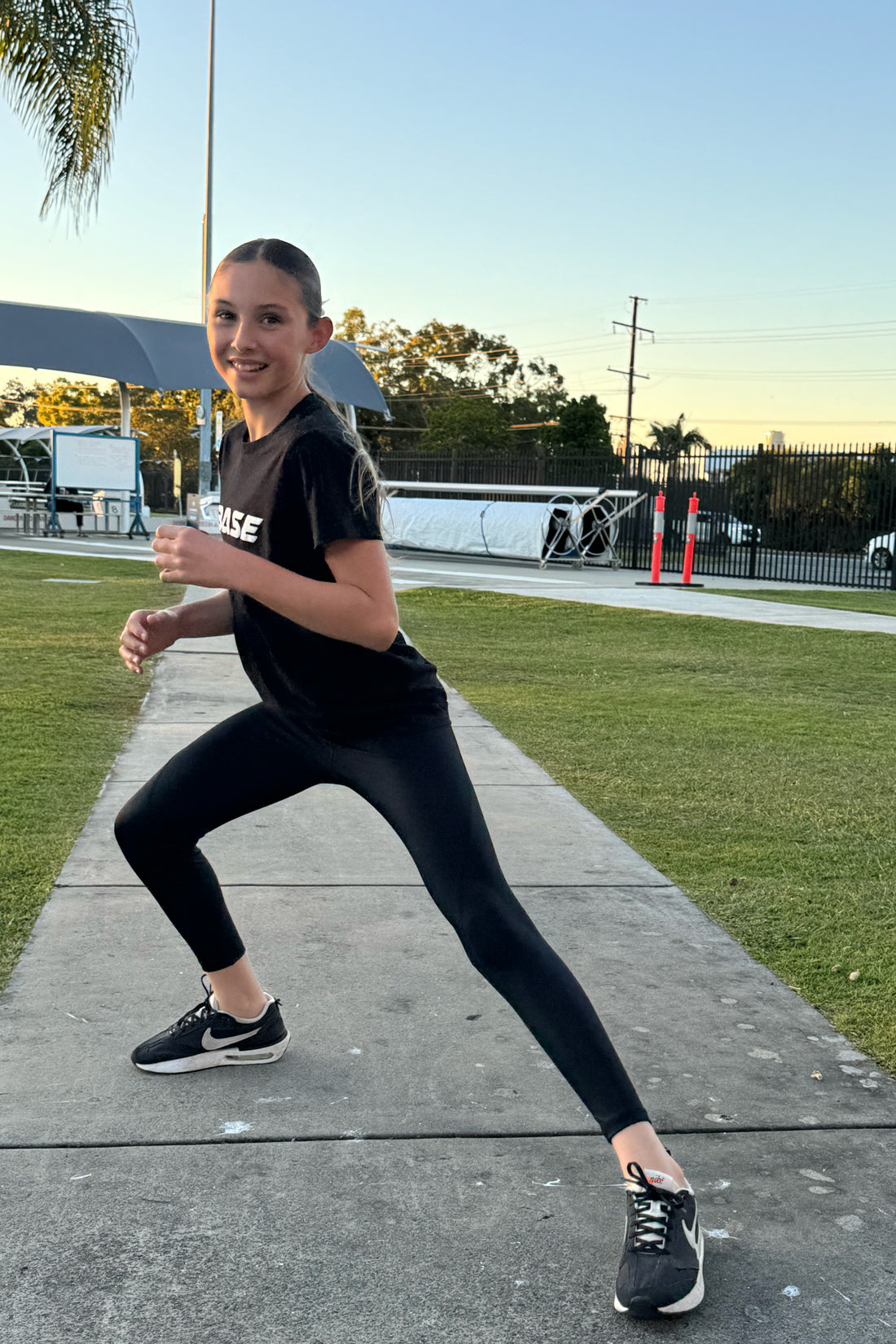Person in black athletic wear standing on a sidewalk with grass and trees in the background