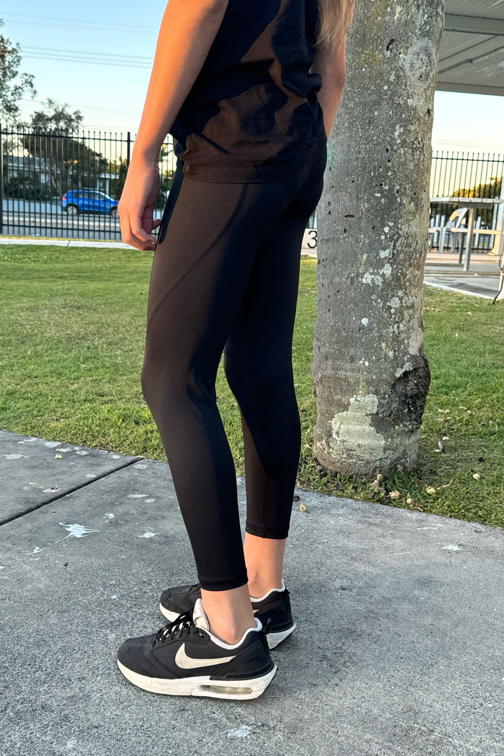 Person wearing black athletic leggings and sneakers standing on a sidewalk with grass and a tree in the background.