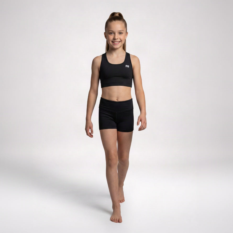 A young girl modeling a black athletic crop top and matching black shorts against a light gray studio background.