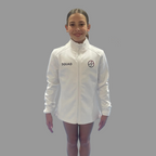 Girl in a white jacket standing on a blue gymnastics floor with other gymnasts in the background.