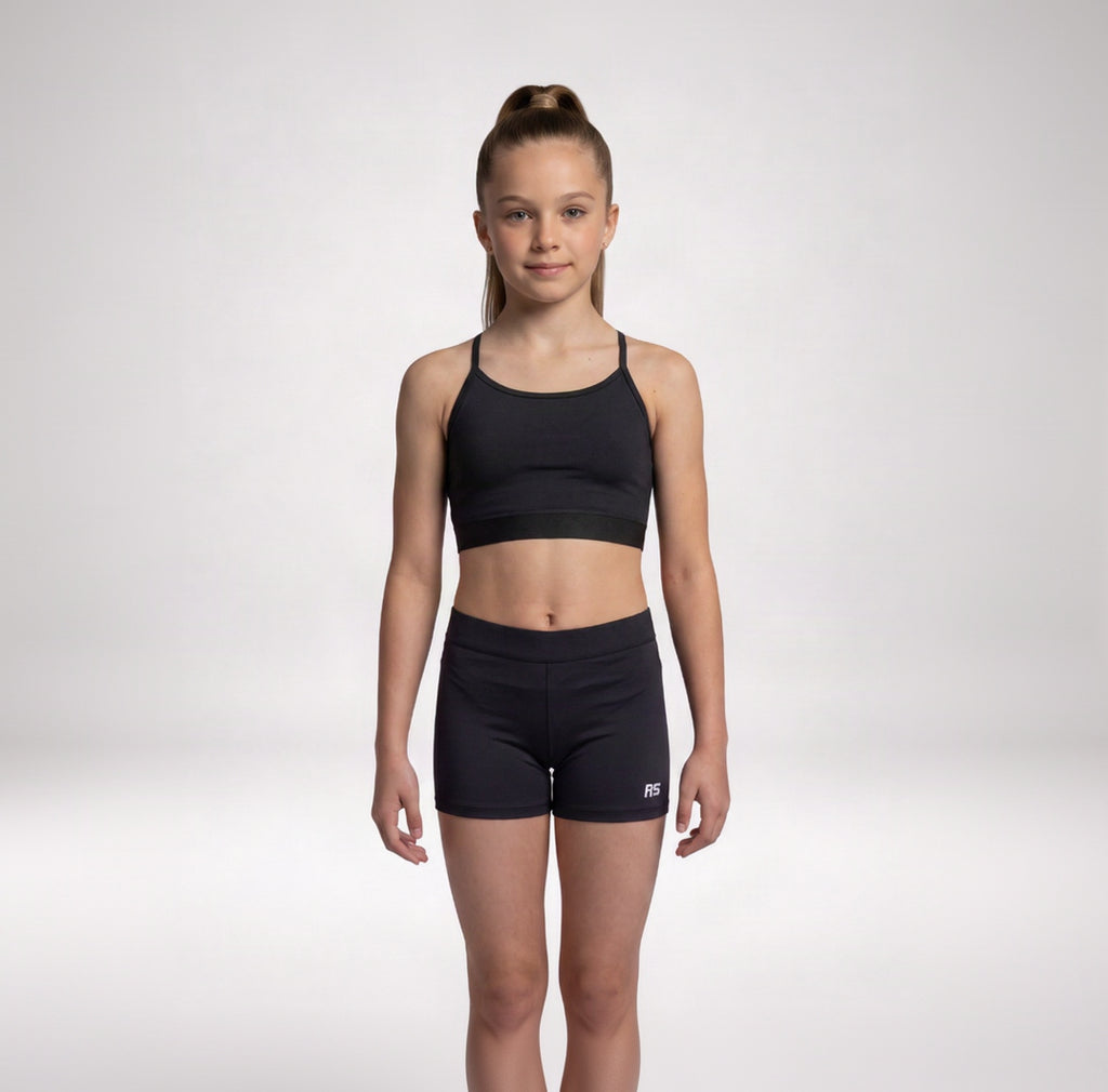 A young girl stands against a light gray studio background wearing a matching black athletic set consisting of a sports bra and fitted shorts. A small white RS logo is visible on the lower right leg of the shorts.